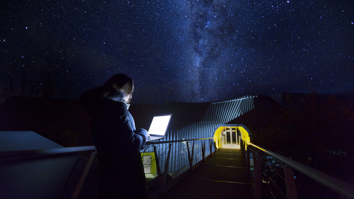 A student stands on a walkway at Mount Stromlo Observatory at night, lit by the light from her laptop screen.
