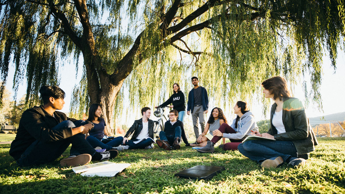 A group of nine ANU students sit in a semi-circle on the grass, under the shade of a huge tree.