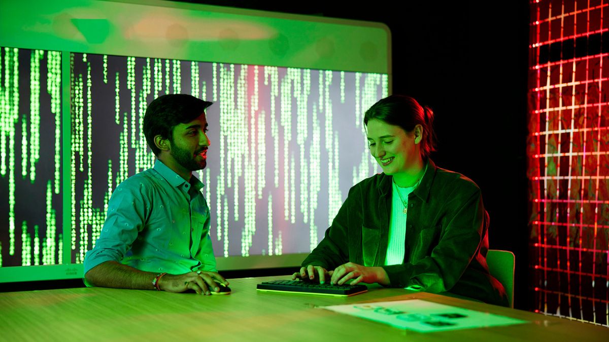 Two people talk while operating a keyboard in a futuristically lit room.