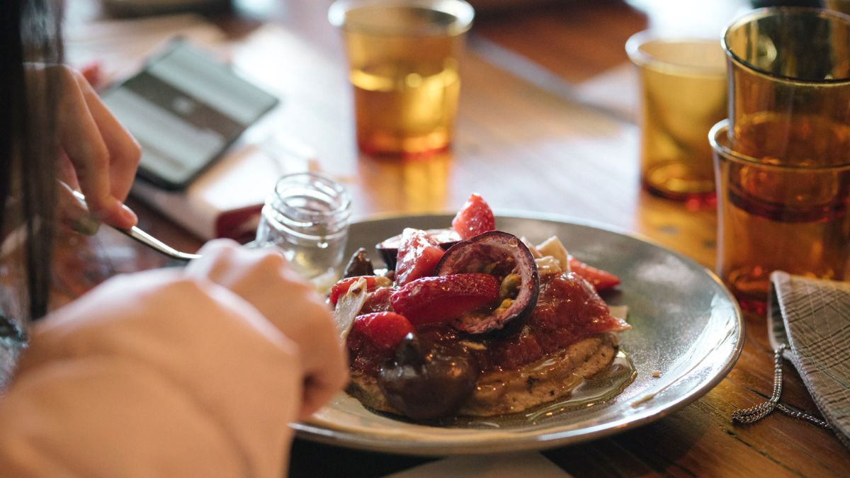 A student eats a plate of vegan breakfast at a Canberra cafe, containing fruit and vegan pancakes. 