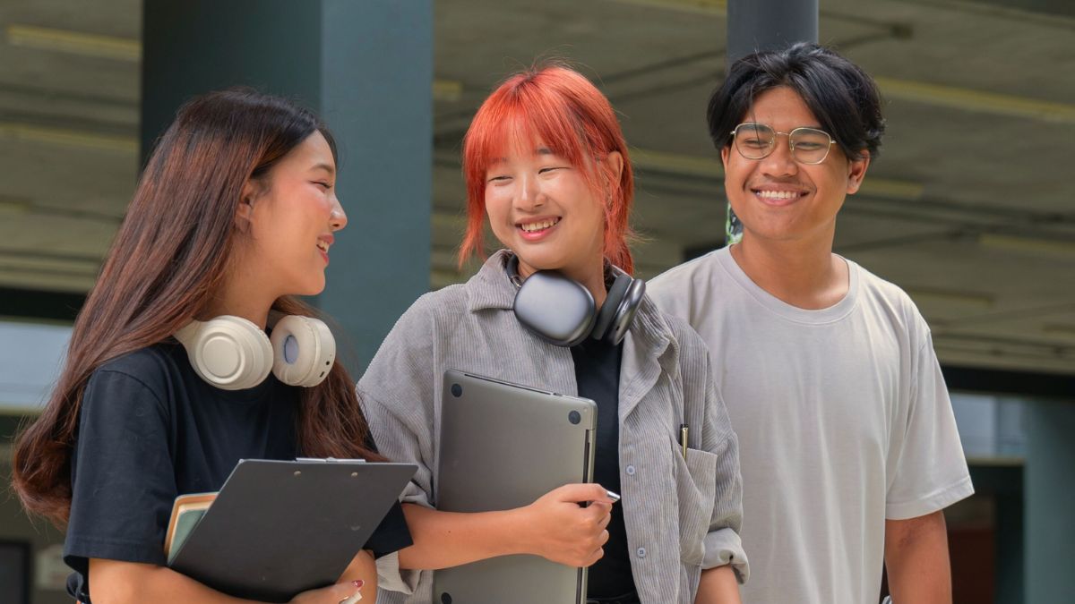 Three university students walking together holding laptops and smiling.