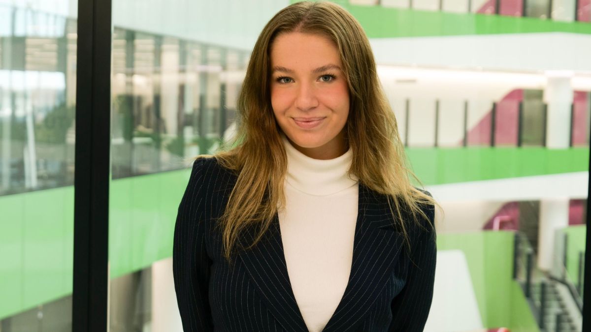 Australian National University student Mikayla stands in front of glass windows inside of a modern campus building.