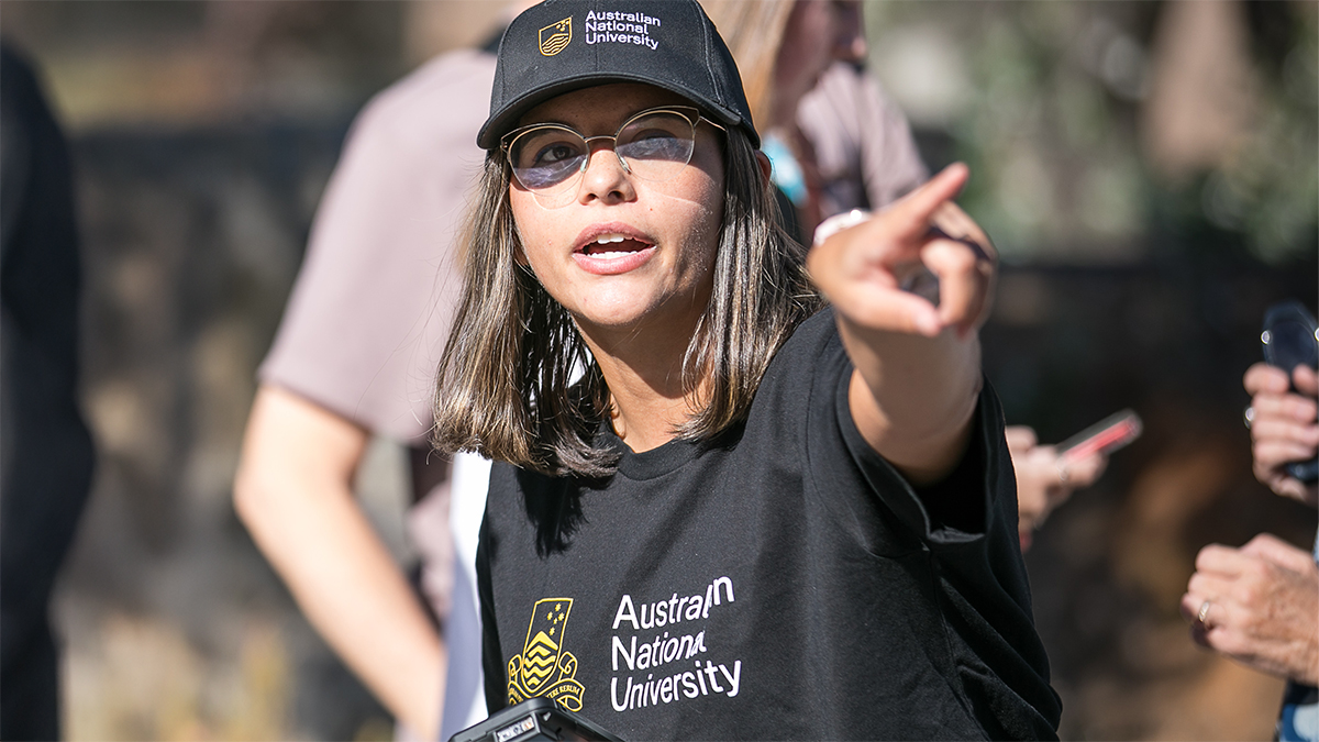 ANU staff member in a university cap and t-shirt leading future students on a campus tour.