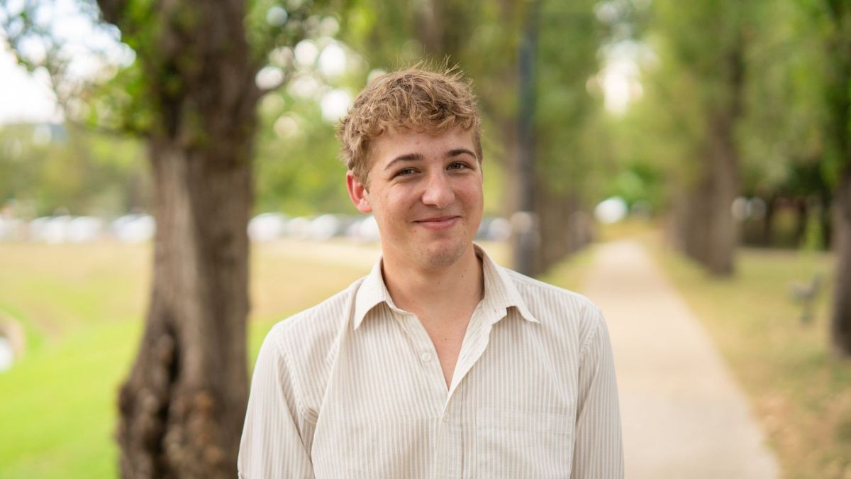 A young man in a light striped shirt smiling on a tree-lined path with greenery in the background.