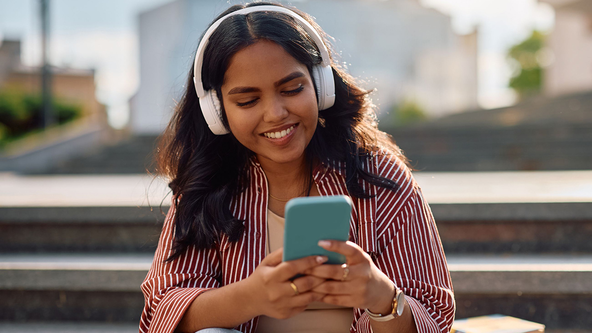 A female student sits on outside stairs with headphones on listening to a podcast, looking at her smartphone. 