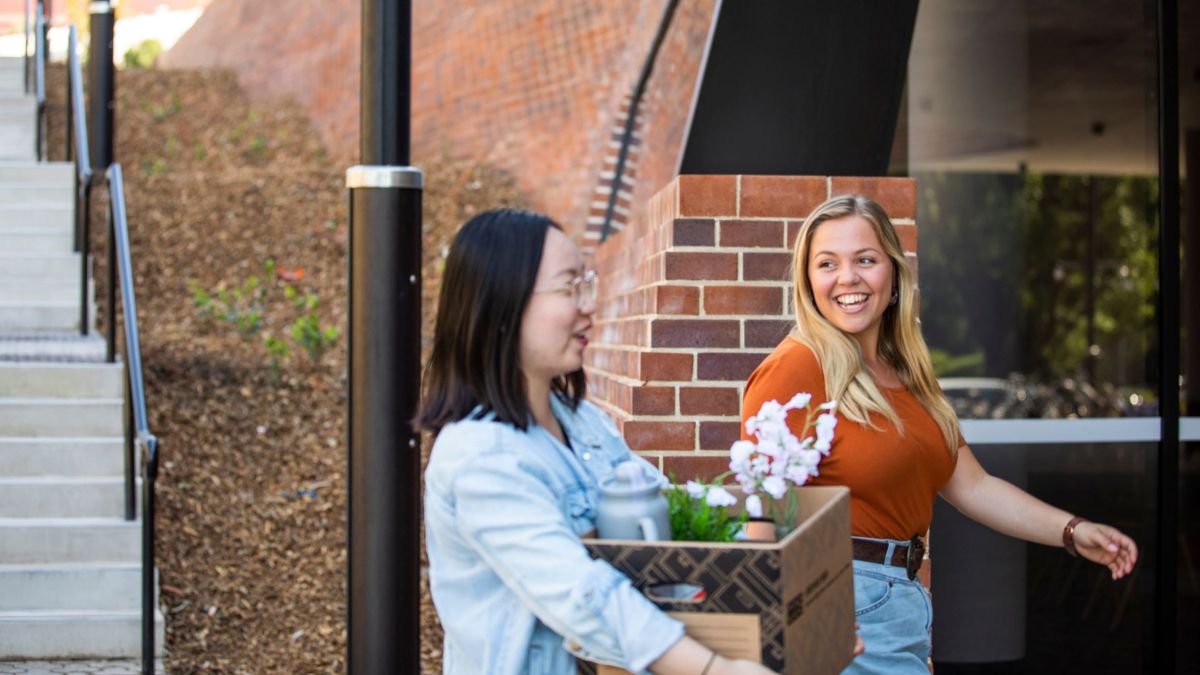 Two women walk into an ANU student residence holding boxes and personal belongings as they move into a room.