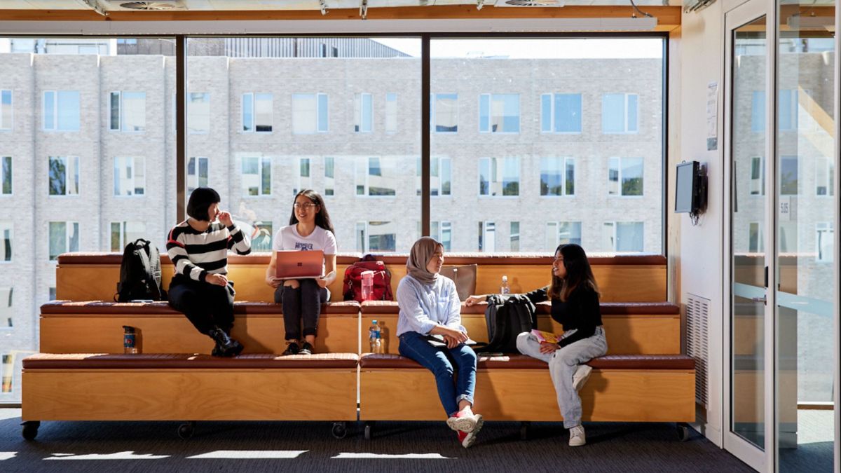Four ANU students sit and chat on tiered seating in the Marie Reay Teaching Centre with large windows behind them.