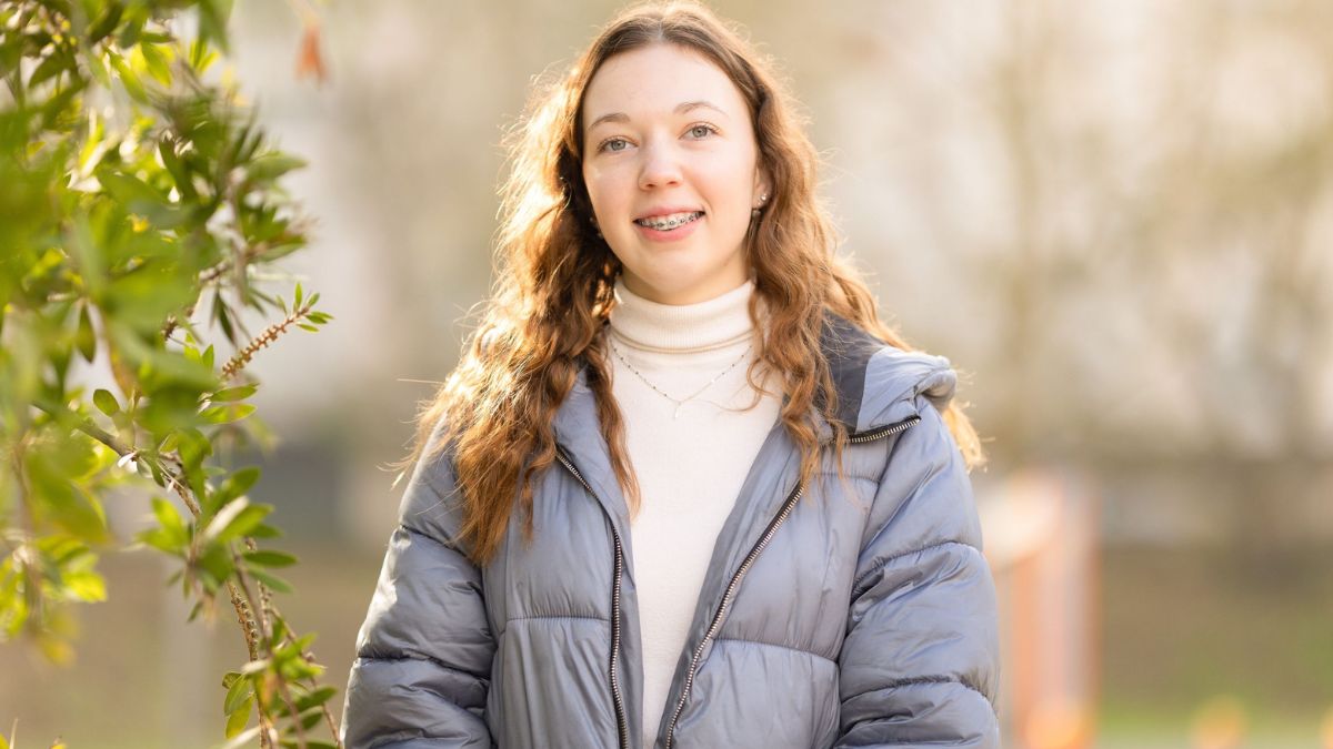 An ANU student smiles for a portrait outdoors, wearing a blue jacket and standing beside green foliage in soft sunlight