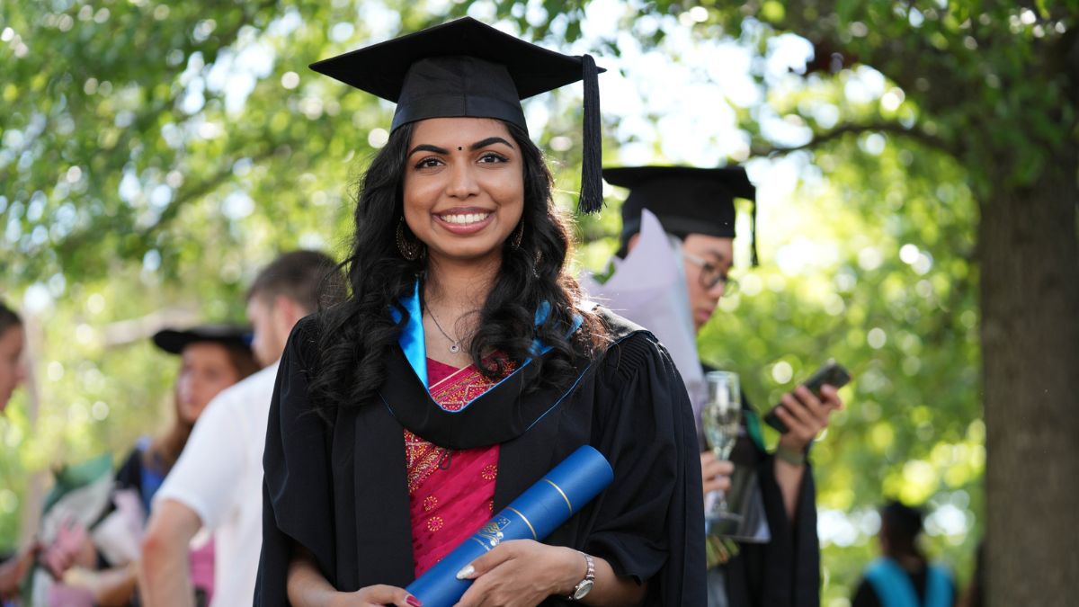 ANU alumni smiles outdoors in academic dress while holding a blue ANU degree scroll, with other graduates gathered in the background.