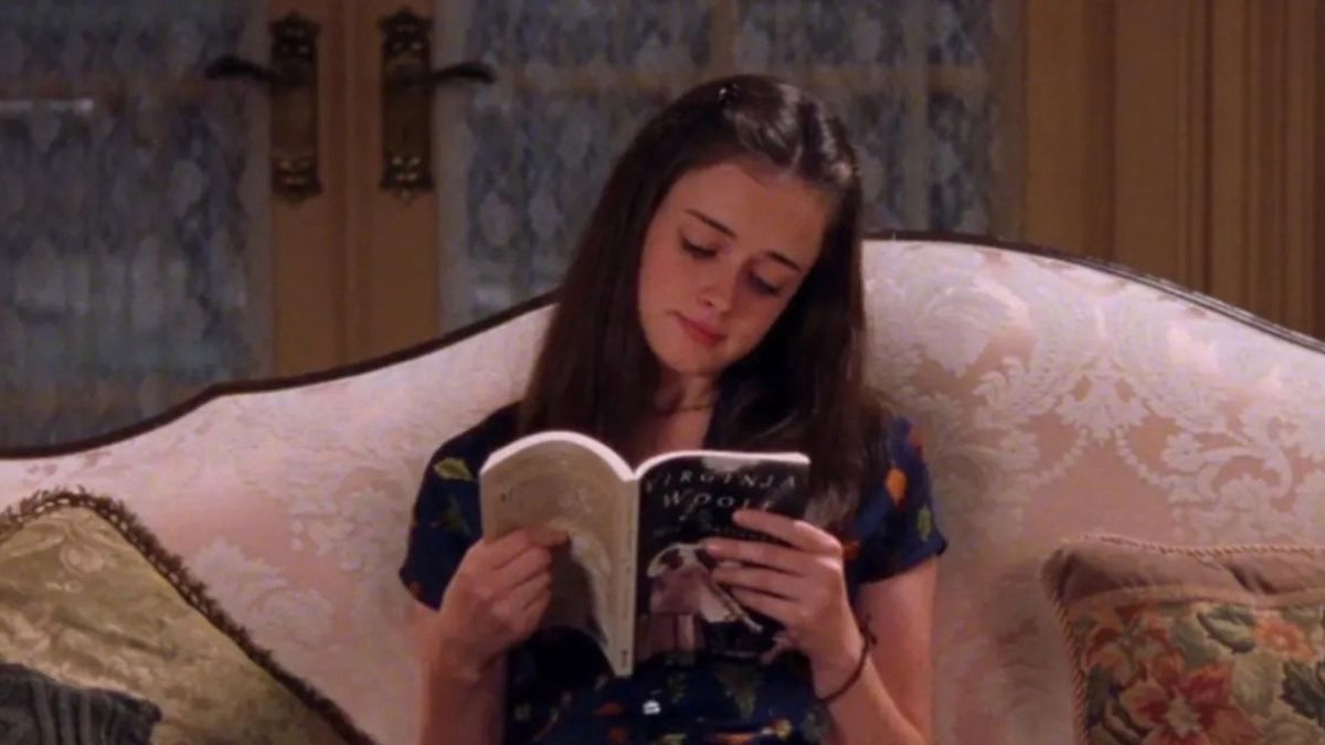 Fictional character Rory Gilmore from the CW show Gilmore Girls sits on a couch reading a book.